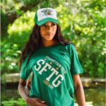 Raven Smith wearing a green SFTG Tee shirt with a Straight from the Go Trucker hat and white tennis skirt in front of a lake and trees.